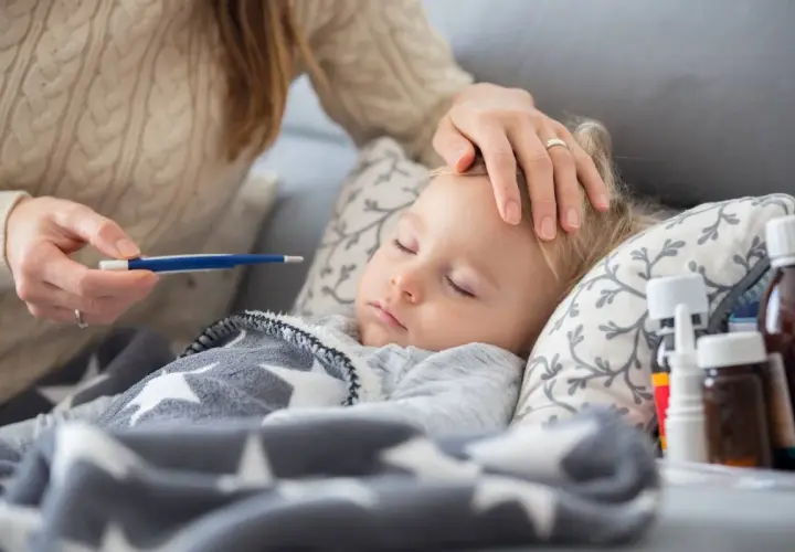 Mama sprawdza temperaturę chorego dziecka. Na stoliku leki, a maluch śpi pod gwiazdkową kołderką. L4 na dziecko.