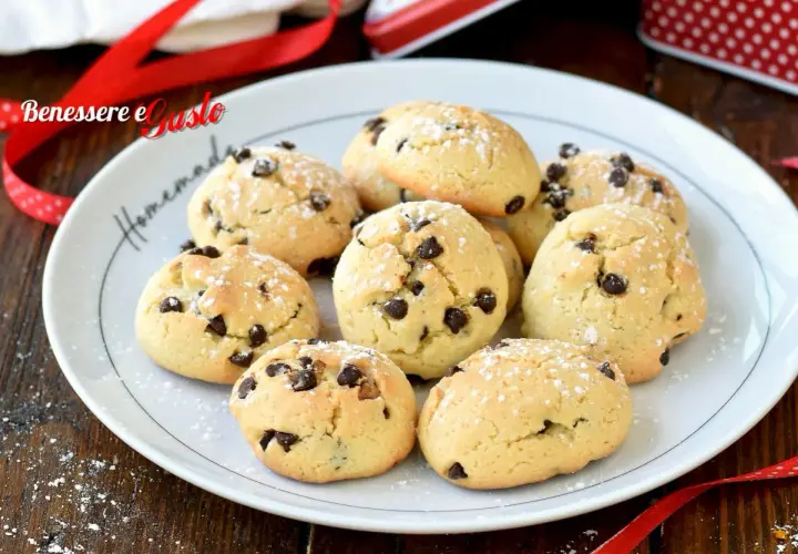 Biscotti con gocce di cioccolato spolverati di zucchero a velo su un piatto bianco, pronti per essere gustati.