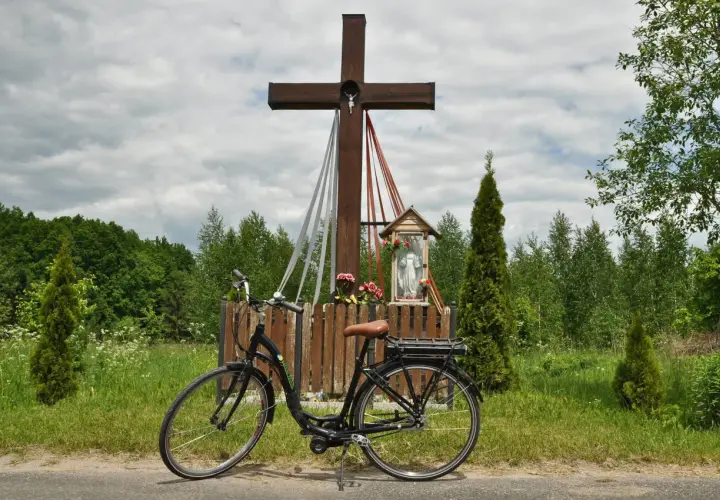 Rower przy kapliczce w Hajnówce, Białowieża. Drewniany krzyż z figurką Chrystusa i Matki Boskiej, otoczony zielenią i polnymi kwiatami.