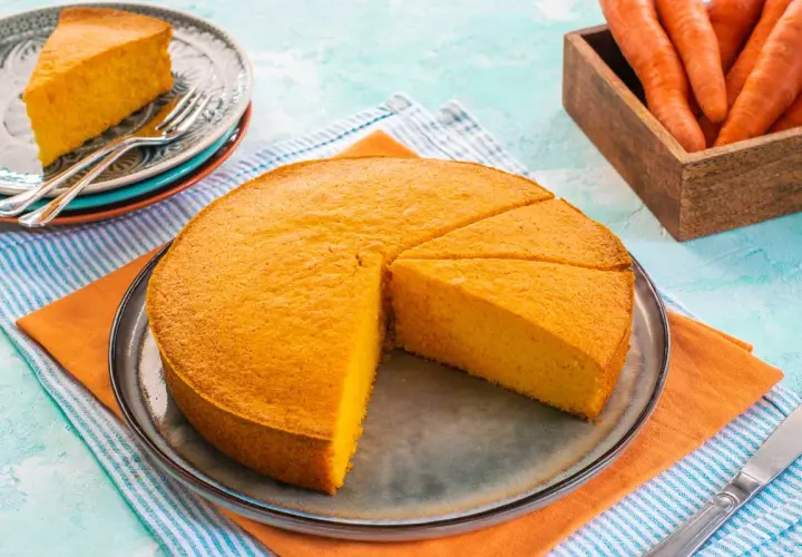 Una deliziosa torta alle carote, soffice e dorata, tagliata a fette su un piatto. Accanto, carote fresche in un cesto di legno.