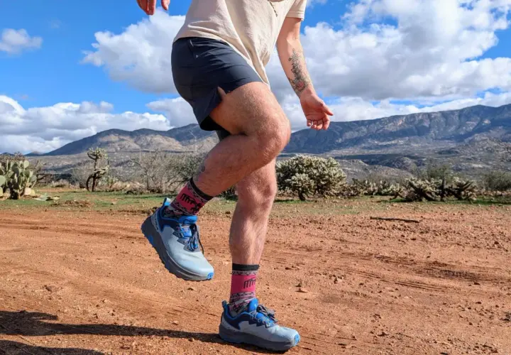 Runner's legs in Altra Timp 6 shoes and patterned socks, mid-stride on a dusty desert trail with mountains in the background.