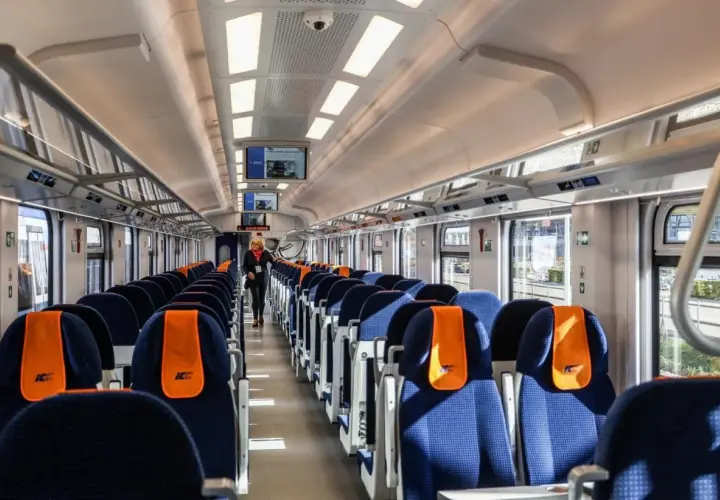Wnętrze wagonu kolejowego z rzędami niebieskich siedzeń z pomarańczowymi zagłówkami.