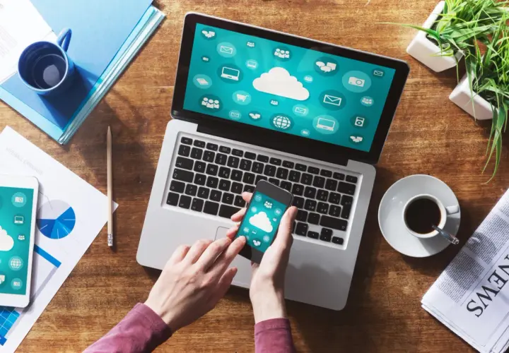 Dłonie trzymające smartfon z ikonami chmury na tle laptopa z podobnymi ikonami.