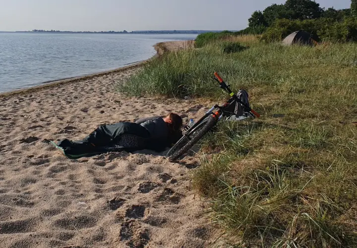 Osoba śpi na plaży obok roweru. W oddali widać wodę i drzewa. To dowód, że można spać na plaży.