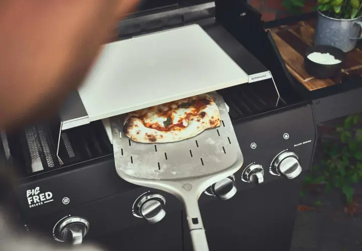 Frisch gebackene Pizza wird mit einem Pizzaschieber aus einem Gasgrill gehoben. Perfekt für den Sommer mit dem Pizza Gasgrill.