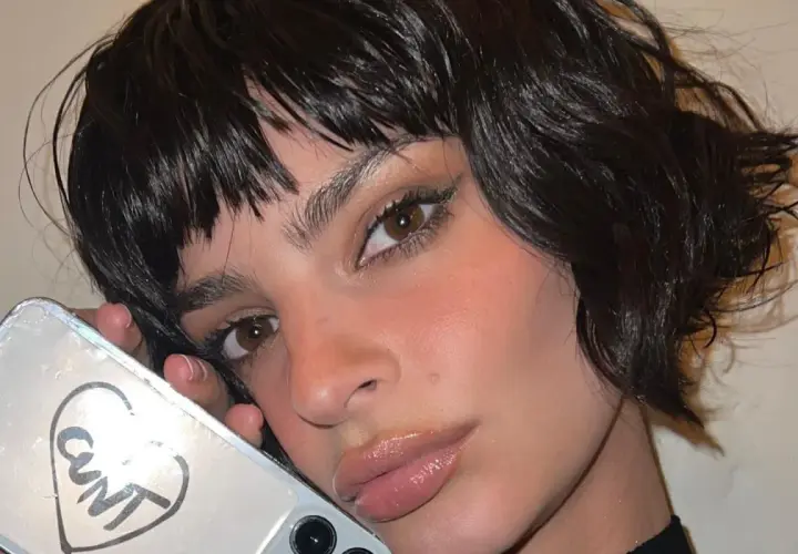 Primo piano di una donna con capelli scuri e umidi che le incorniciano il viso. Tiene in mano un telefono con un disegno a cuore e la scritta "CUNT".