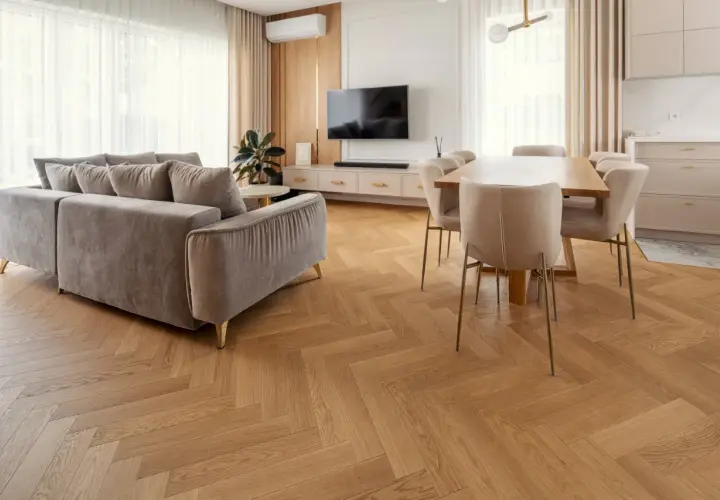 Eleganckie wnętrze z szarą kanapą i jadalnią. Wzrok przyciąga piękna podłogówka ułożona we wzór jodełki.