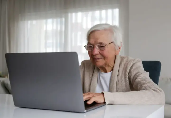 Uśmiechnięta starsza pani z okularami pracuje na laptopie, planując swoją emeryturę bez podatku.