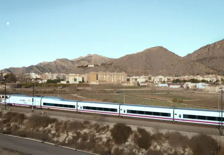 Tren AVE de Renfe, un moderno tren blanco con detalles morados, atraviesa un paisaje rural con montañas y edificios al fondo.