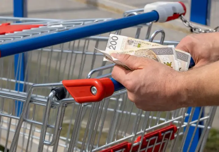 Ręce trzymają banknoty 200 zł przy wózku sklepowym. Kto zyskuje na inflacji, gdy ceny rosną?