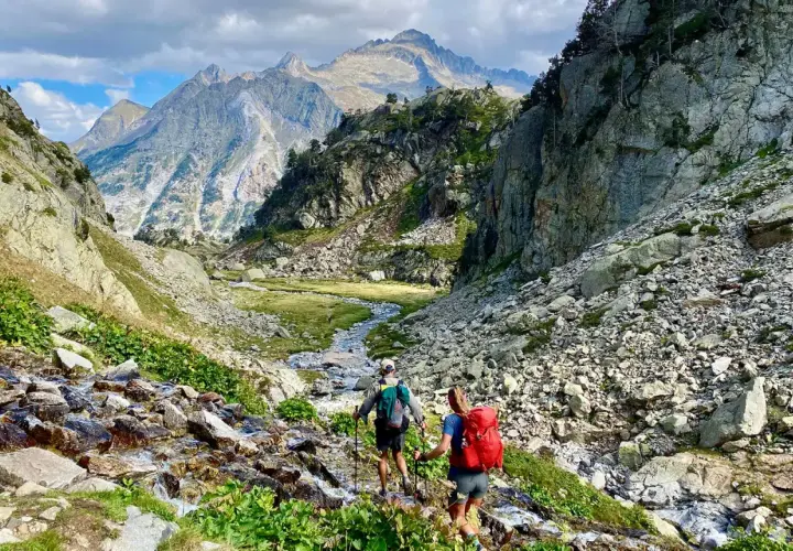Hikers ascend a rocky path alongside a stream, with majestic mountains in the distance, on the Haute Route Pyrenees.