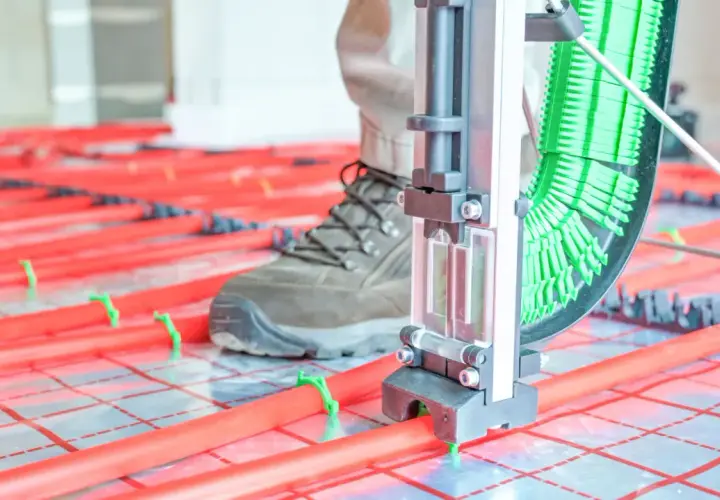 Instalator z narzędziem układa rury do ogrzewania podłogowego. Czerwone rurki na zielonych klipsach tworzą siatkę.