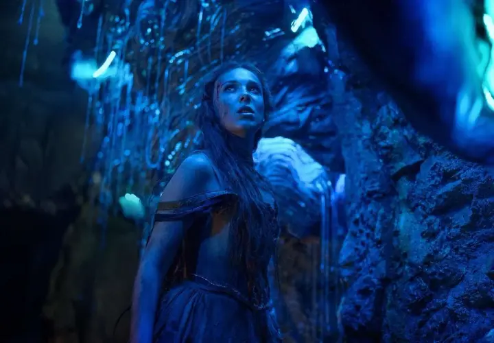 Una giovane donna con lunghi capelli scuri, vestita con abiti blu, guarda verso l'alto in una grotta illuminata da luci blu. La sua espressione è di stupore e meraviglia, quasi come se fosse una damigella in pericolo in un mondo alieno. La recensione d...