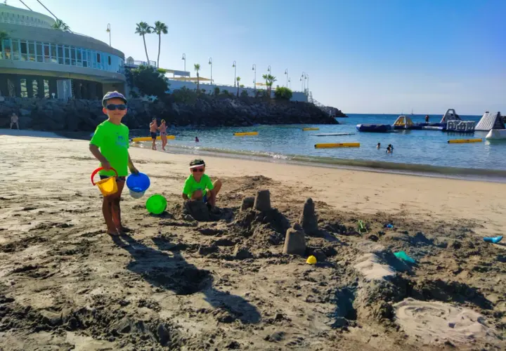 Radosne **teneryfa z dziećmi** na plaży. Dwóch chłopców buduje zamki z piasku, a w tle widać wodne atrakcje.
