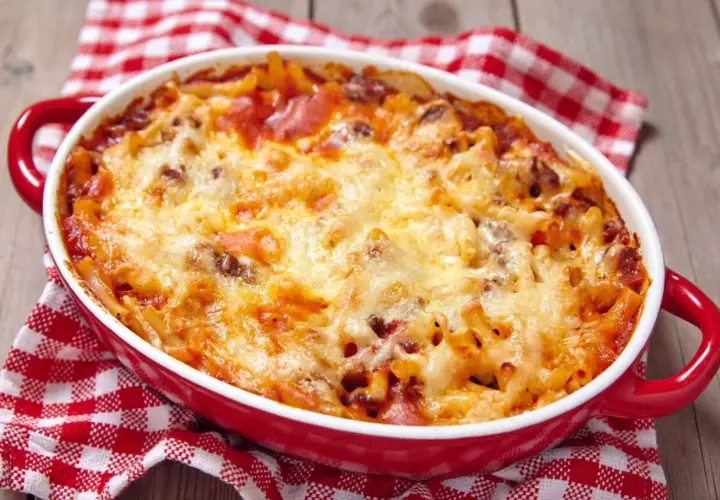 Zapiekanka makaronowa z mięsem mielonym i roztopionym serem w czerwonym naczyniu, na tle kratkowanej serwetki.