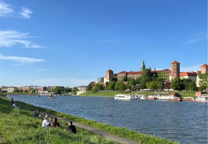 Słoneczny dzień nad Wisłą, ludzie odpoczywają na trawie, a w tle majaczy Wawel – symbol starego miasta Krakowa.