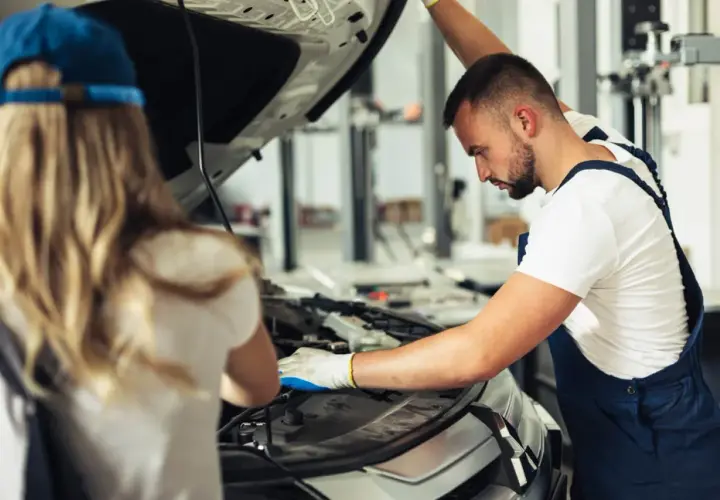 Mechanik z rękawiczkami bada silnik, szukając przyczyny usterki systemu zarządzania silnikiem. Obok stoi kobieta w czapce.