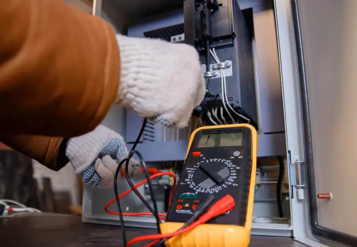 Elektryk w rękawiczkach sprawdza połączenia w szafie sterowniczej za pomocą żółtego multimetru. Pokazuje, jak używać multimetru do pomiarów.