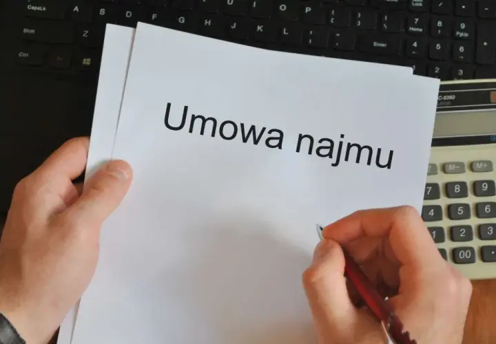 Ręka pisze długopisem umowę najmu. Obok leży kalkulator i klawiatura, gotowe do pracy dla wynajmującego.