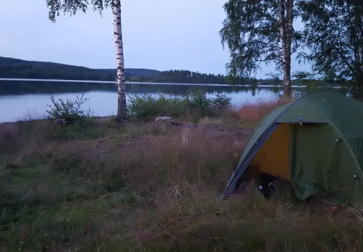 En grön campingtält står vid en sjö, med en skogklädd kulle i bakgrunden. En perfekt plats för äventyr längs 7 torpsleden.