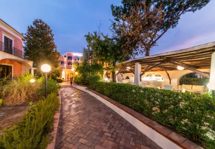 Sentiero in mattoni che porta a un bar all'aperto illuminato, con edifici color pastello sullo sfondo. Un'atmosfera da sogno al Valentino Ischia.