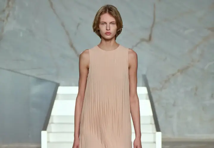 Modelo con vestido plisado color melocotón, en un escenario minimalista con escaleras blancas y fondo de mármol.
