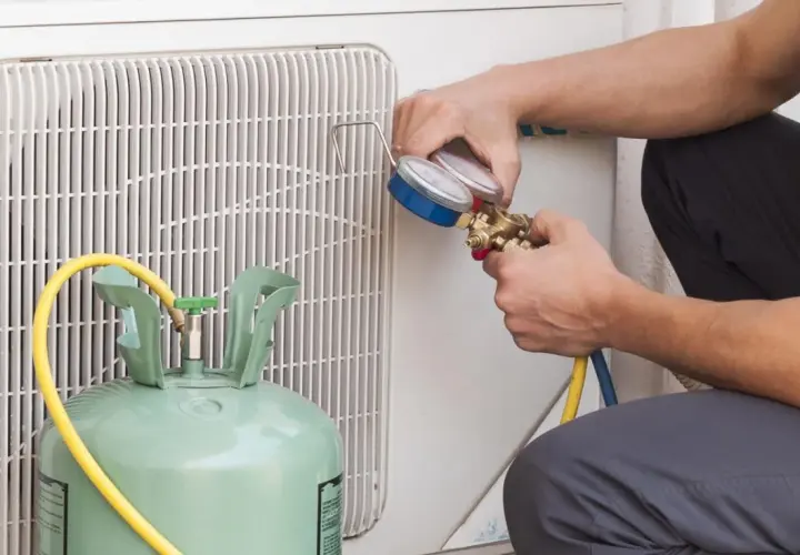 Técnico revisa manómetro para detectar fuga aire acondicionado. Cilindro de gas refrigerante y unidad exterior visibles.
