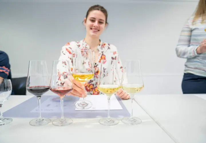 Eine Frau mit Blumenmuster-Bluse verkostet Wein, ein Schritt auf dem Weg, Sommelier zu werden. Mehrere Gläser mit Rot-, Rosé- und Weißwein stehen auf dem Tisch.