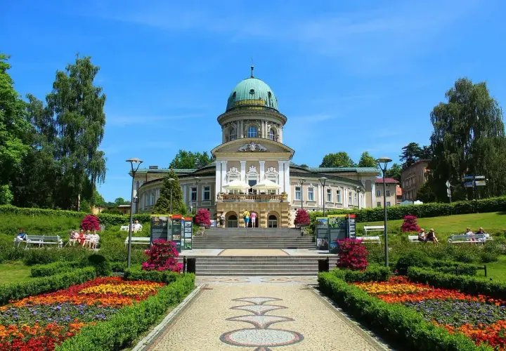 Piękny budynek uzdrowiskowy z kopułą, otoczony kwitnącym ogrodem i alejkami. Idealne miejsce na relaks i spacer, jakby czas się tu zatrzymał.