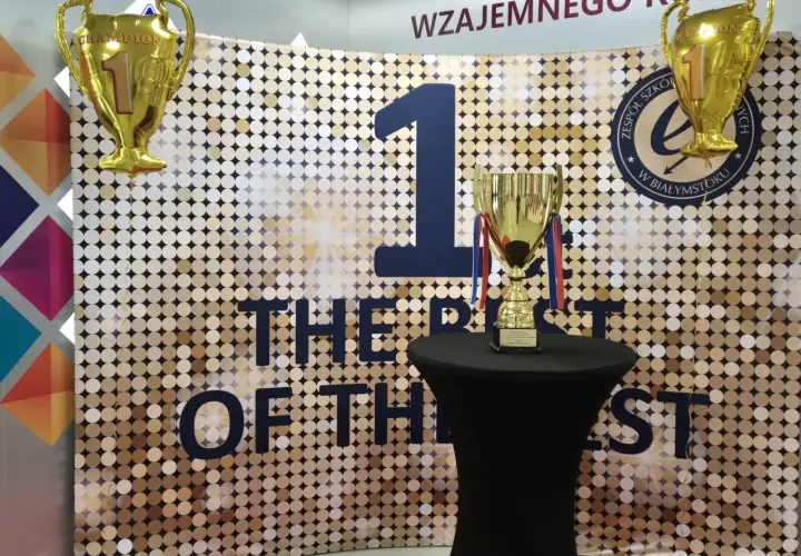 Złote puchary i balon w kształcie pucharu na tle błyszczącej ścianki z napisem "THE BEST OF THE BEST".