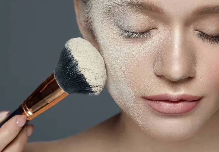 Applicazione di cipria per il baking trucco su un viso femminile. Un pennello grande raccoglie la polvere per un finish impeccabile.