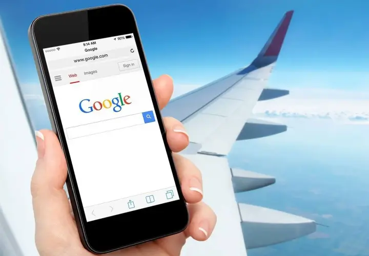 Dłoń trzyma smartfon z otwartą wyszukiwarką Google, na tle skrzydła samolotu i nieba. Czy w samolocie można mieć internet? Tak, można!