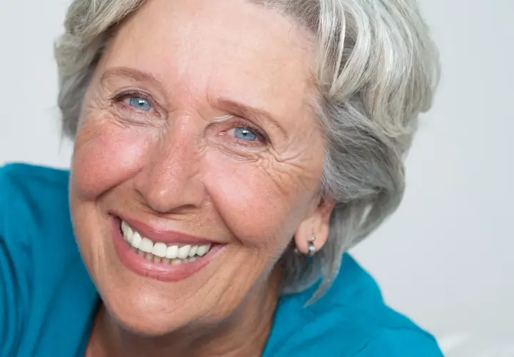 Donna anziana con capelli grigi e occhi azzurri, sorride mostrando le rughe intorno alla bocca, segno di una vita vissuta con gioia.