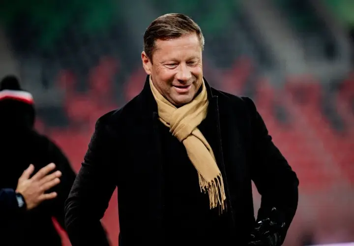 Uśmiechnięty Dariusz Banasik w czarnym płaszczu i beżowym szaliku, na tle trybun stadionu.