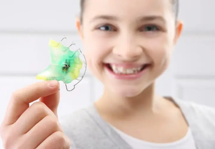 Niña sonriente muestra un retenedor dental verde y amarillo, listo para limpiar.
