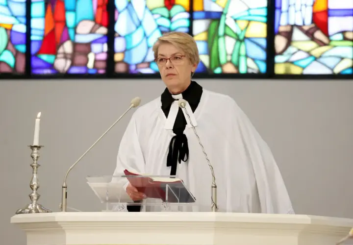 Kobieta w stroju duchownego, z białą narzutą i czarną kryzą, stoi przy pulpicie. To pastor, który przygotowuje się do wygłoszenia kazania.