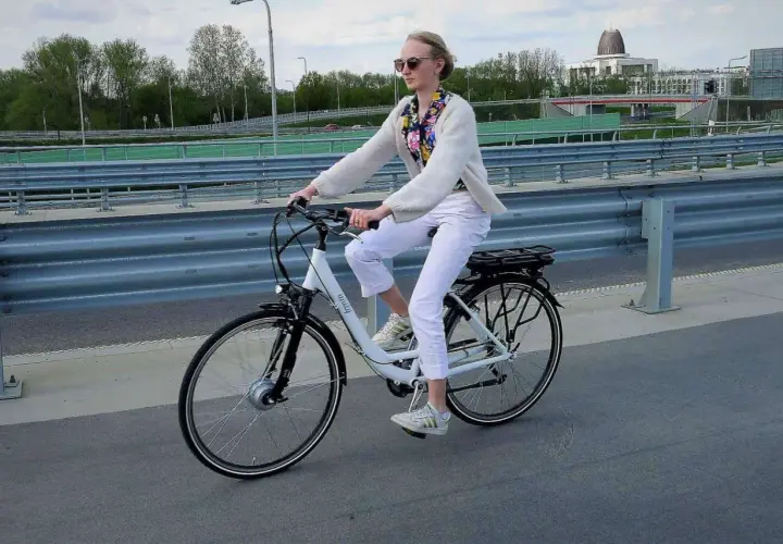 Kobieta w białych spodniach i sweterku jedzie na rowerze elektrycznym. W tle most i budynki.