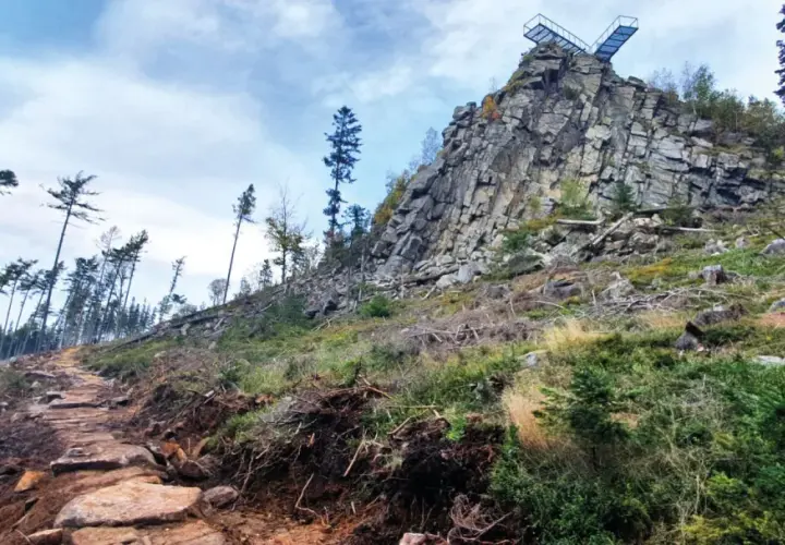 Kamienista ścieżka prowadzi ku skalistemu szczytowi z platformą widokową. W oddali widać las, a krajobraz przypomina ten z **Trójaka w Lądku Zdroju**.