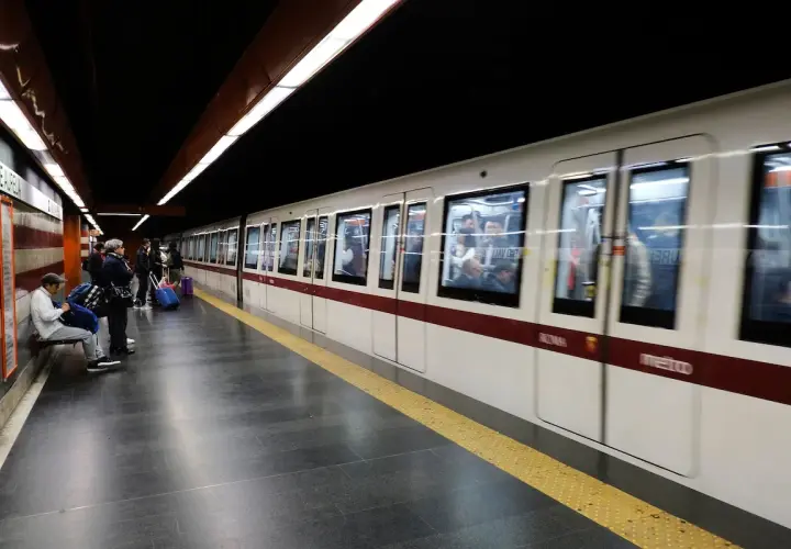Des voyageurs attendent sur le quai du **metro a Rome**. Un train blanc et bordeaux arrive, reflétant les lumières du tunnel.
