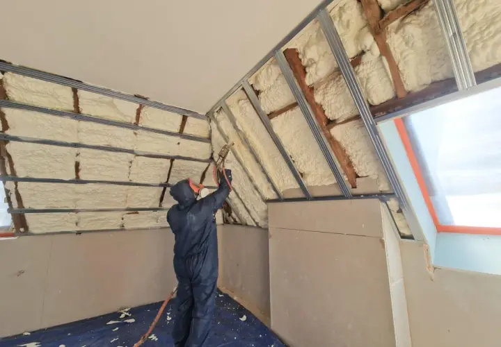 Un ouvrier en combinaison projette de la mousse polyuréthane pour isoler un grenier. L'isolant blanc recouvre les poutres et les murs.