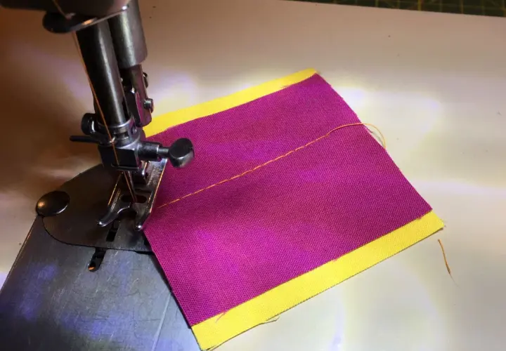 Close-up of a sewing machine needle and foot over purple fabric with a yellow border, demonstrating how to thread a sewing machine.