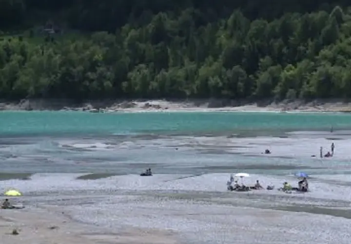 Persone rilassate sulla spiaggia del lago di Barcis, con acque turchesi e boschi sullo sfondo.