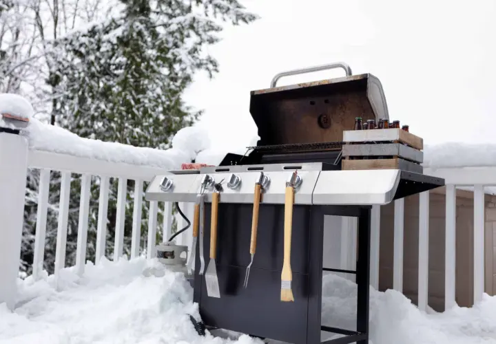 Un barbecue d'hiver prêt à l'emploi, avec des bouteilles de bière dans une caisse en bois et des ustensiles. La neige recouvre la terrasse et la balustrade.