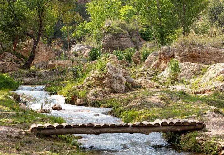 Un puente rústico cruza un arroyo en un paisaje rocoso y verde, evocando la serenidad de una cascada en Chelva.