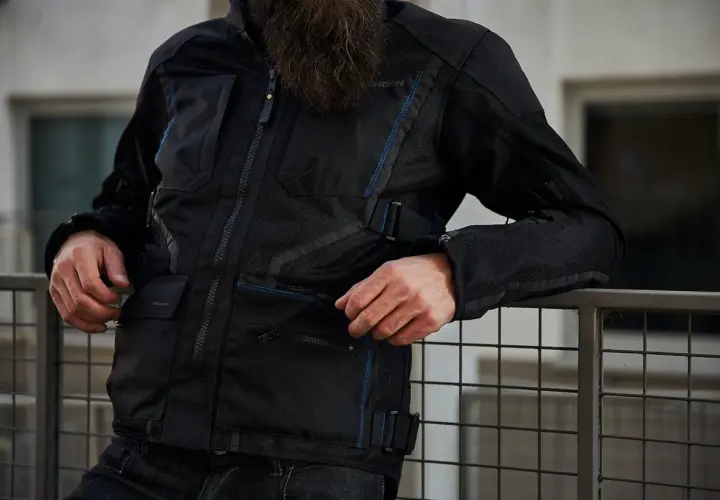 Mężczyzna w czarnej kurtce motocyklowej Rebelhorn Scandal II z niebieskimi akcentami, oparty o metalową barierkę.