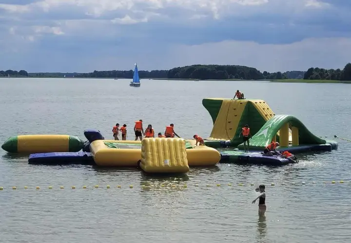 Dzieci w kamizelkach ratunkowych bawią się na dmuchanych zjeżdżalniach i torach przeszkód na jeziorze. Idealne miejsce, gdzie na Mazury z dziećmi!
