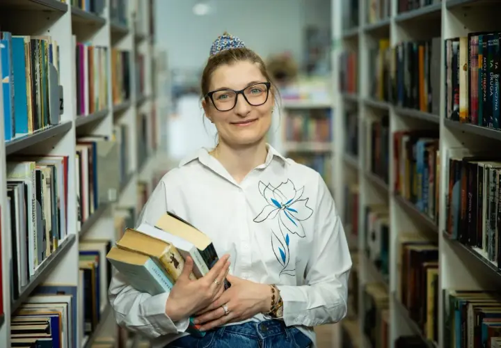 Bibliotekarka z okularami trzyma stos książek. W tle regały pełne literatury. Czy wakat co to znaczy?