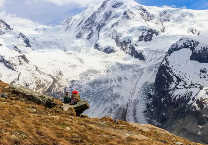 Two hikers rest on a rocky outcrop, gazing at the majestic, snow-covered peaks of the tour de Monte Rosa.