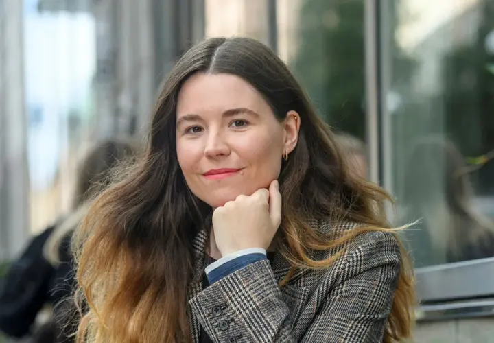 Johanna Hedman, med sitt långa, vågiga hår och en rutig kavaj, ler mot betraktaren.