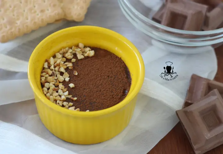 Budino al cioccolato in ciotola gialla, cosparso di cacao e nocciole tritate. Accanto, biscotti e cioccolato.